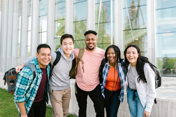 A group of five smiling friends, each wearing casual clothing and backpacks, stand close together outside a modern building.